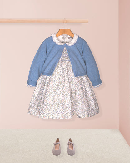 The Fiorella Belina dress styled with the Serena Cardigan in a soft blue knit and paired with navy blue Mary Jane shoes, displayed against a pastel pink backdrop.