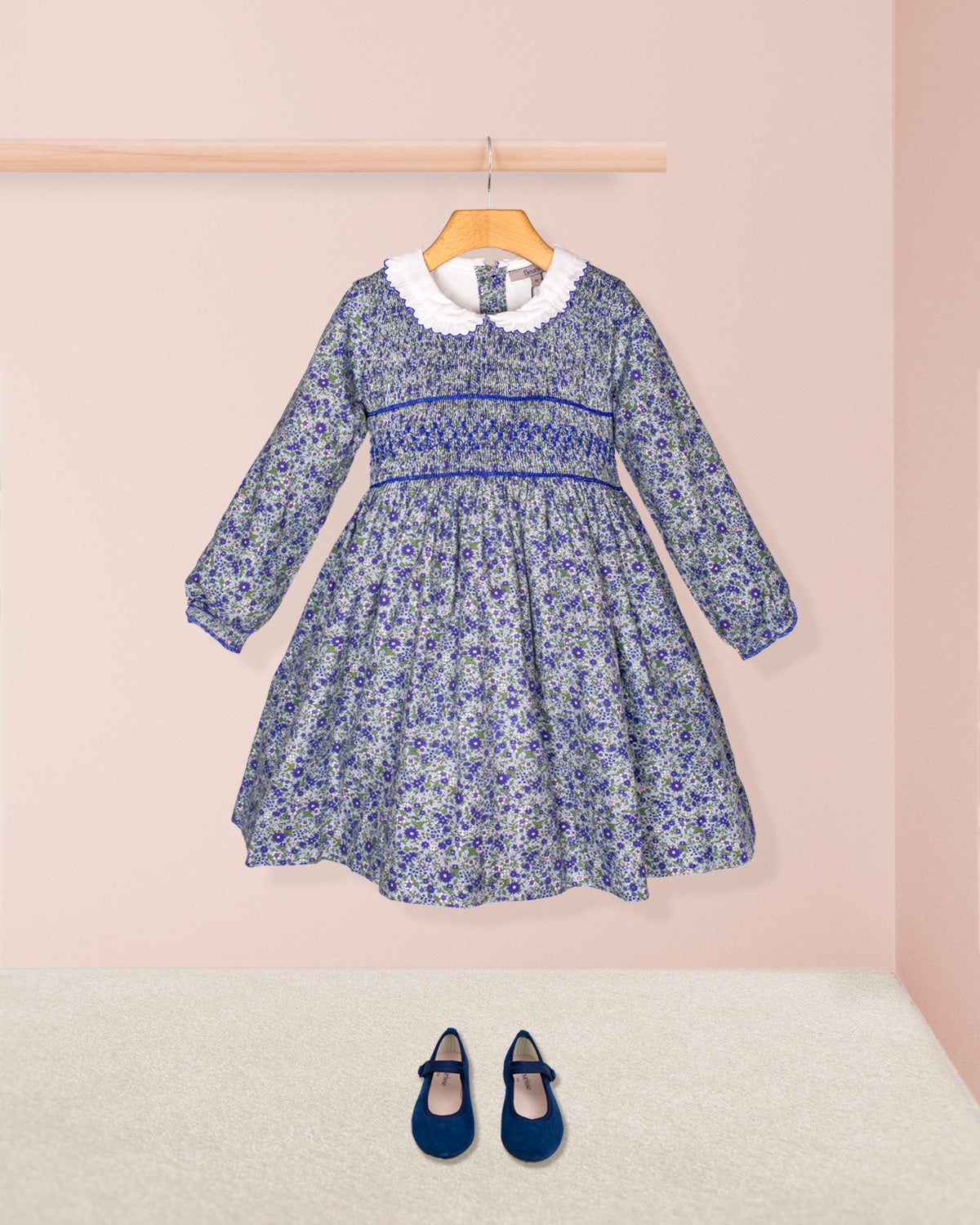 The Fiorella Mini Blue Floral Twill Smocked Dress styled with navy Mary Jane shoes, shown against a blush pink background to highlight the full outfit in a standalone look