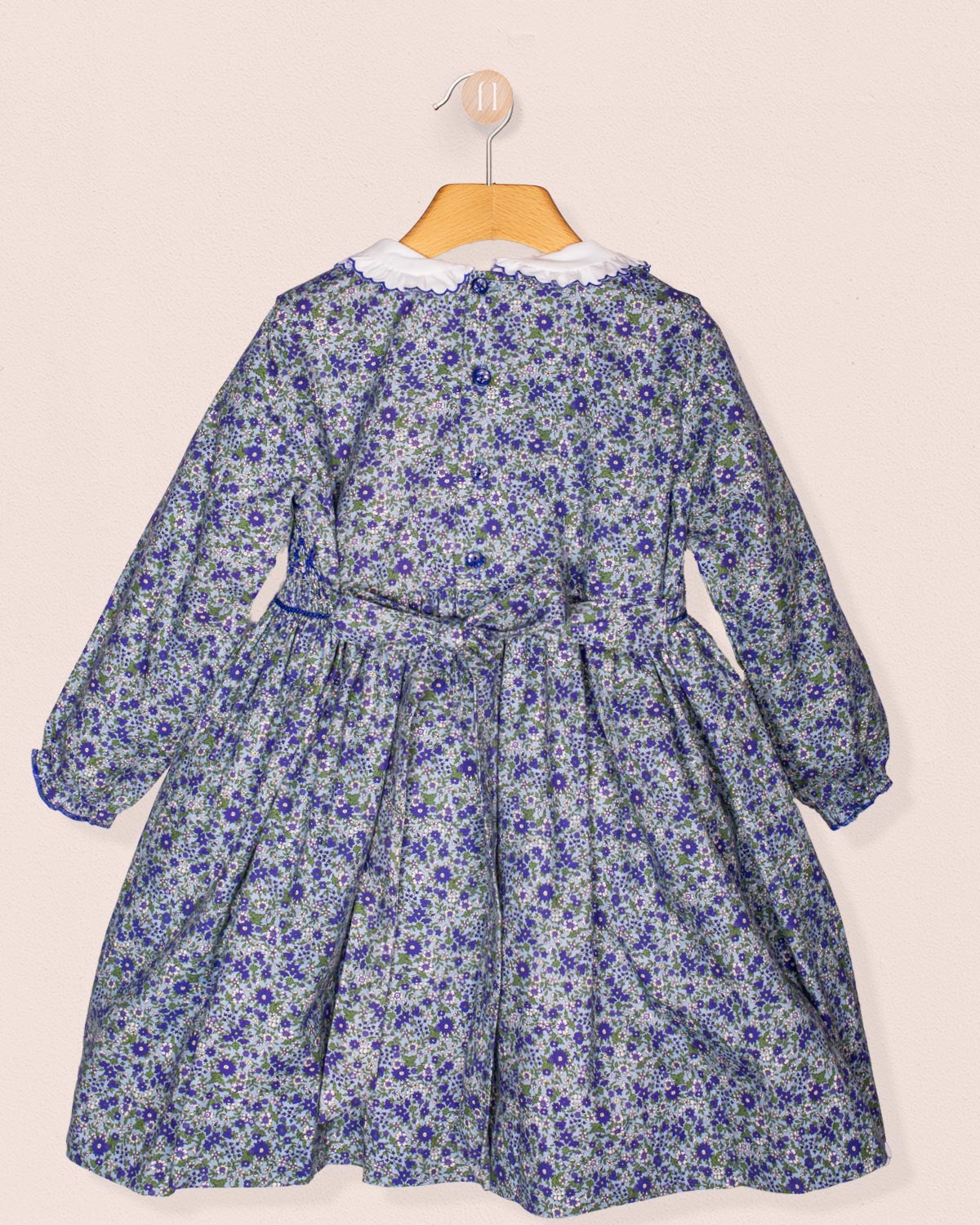 Back view of the Fiorella dress hanging on a wooden hanger, highlighting the tie-back sash and gathered waist against a soft beige background.