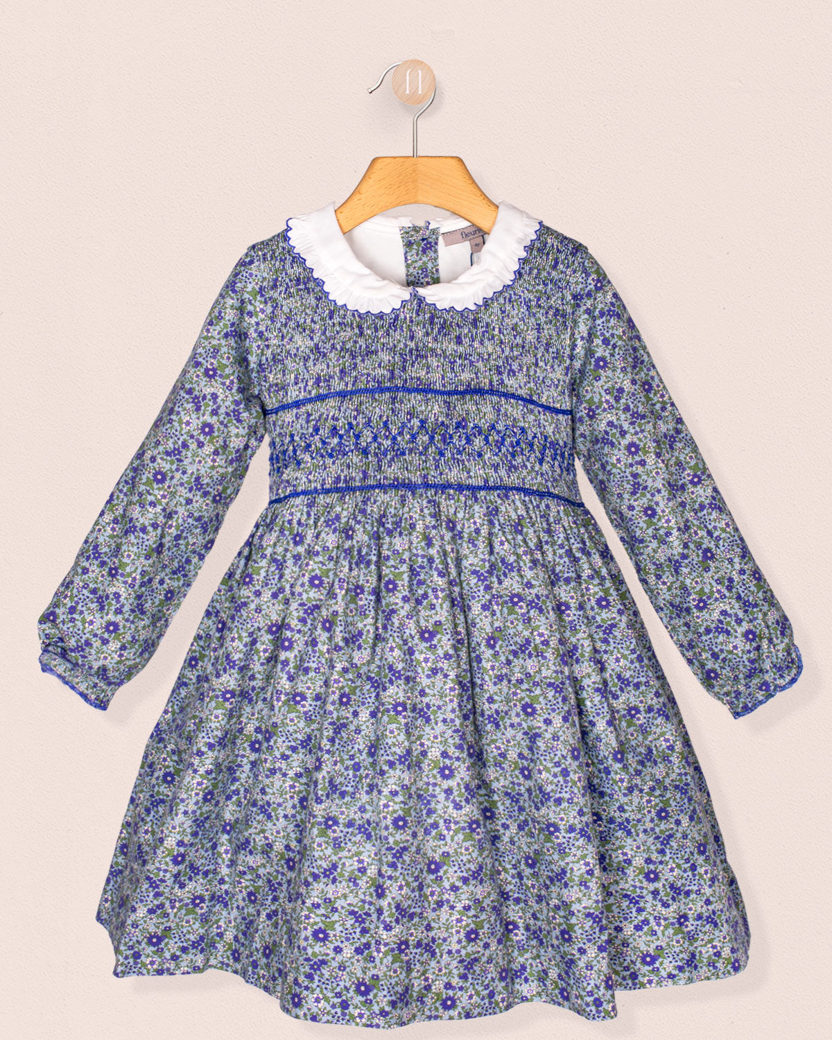 A front-facing view of the Fiorella Belina dress, featuring a mini blue floral print, hand-smocked bodice, scalloped white collar, and long puffed sleeves with elastic cuffs. The dress is displayed on a wooden hanger against a soft pink wall.