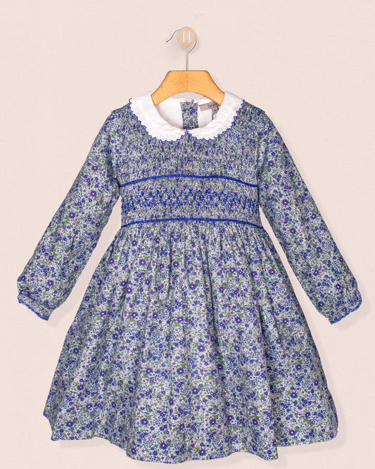 A front-facing view of the Fiorella Belina dress, featuring a mini blue floral print, hand-smocked bodice, scalloped white collar, and long puffed sleeves with elastic cuffs. The dress is displayed on a wooden hanger against a soft pink wall.
