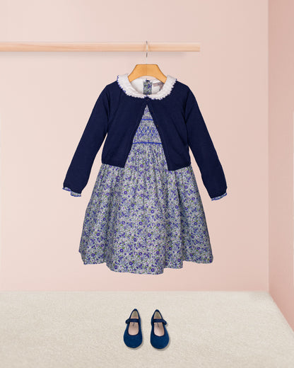 The Fiorella Belina dress styled with the Serena Cardigan in a navy knit and paired with navy blue suede Mary Jane shoes, displayed against a pastel pink backdrop.