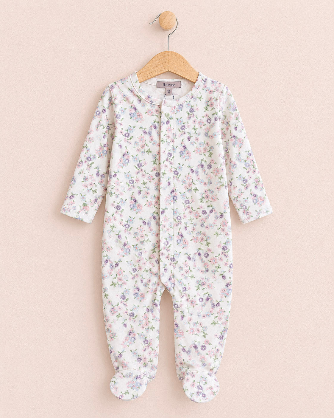 A baby footed sleeper with long sleeves and an all-over small floral print, hanging on a wooden hanger against a solid pale pink background.