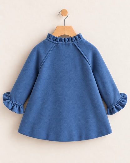Backside of a blue coat on hanger against a neutral tan background, withruffled neckline & sleeve cuffs at the wrist.