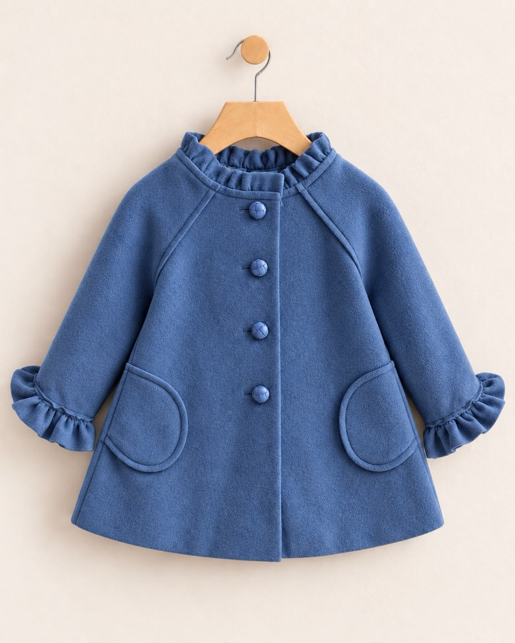Frontside of a blue coat on hanger against a neutral tan background, with 4 vertical buttons, 2 pockets and ruffled neckline & sleeve cuffs at the wrist.