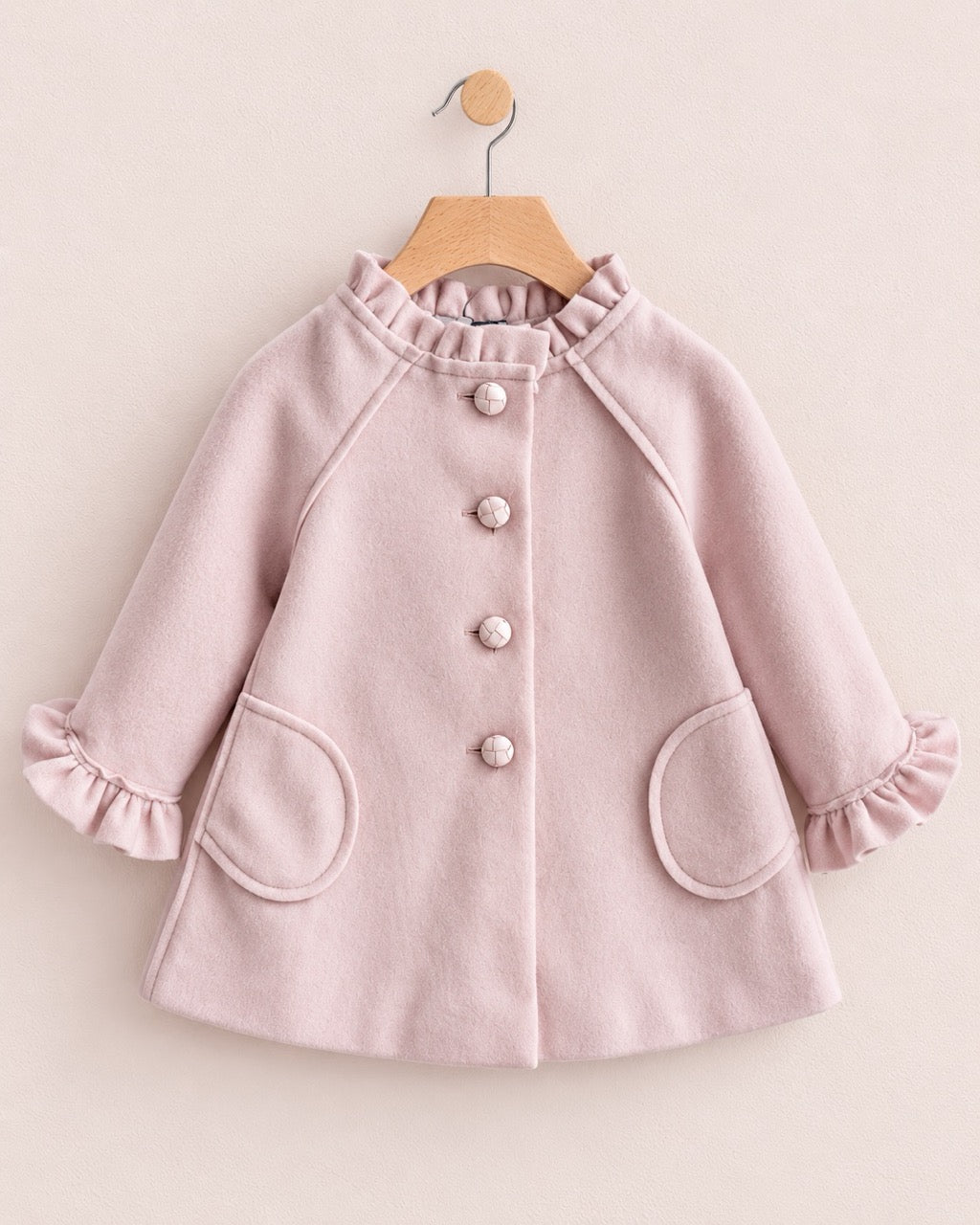 Light pink colored coat with 4 buttons and 2 oval shaped pockets, on a hanger against a muted pink background