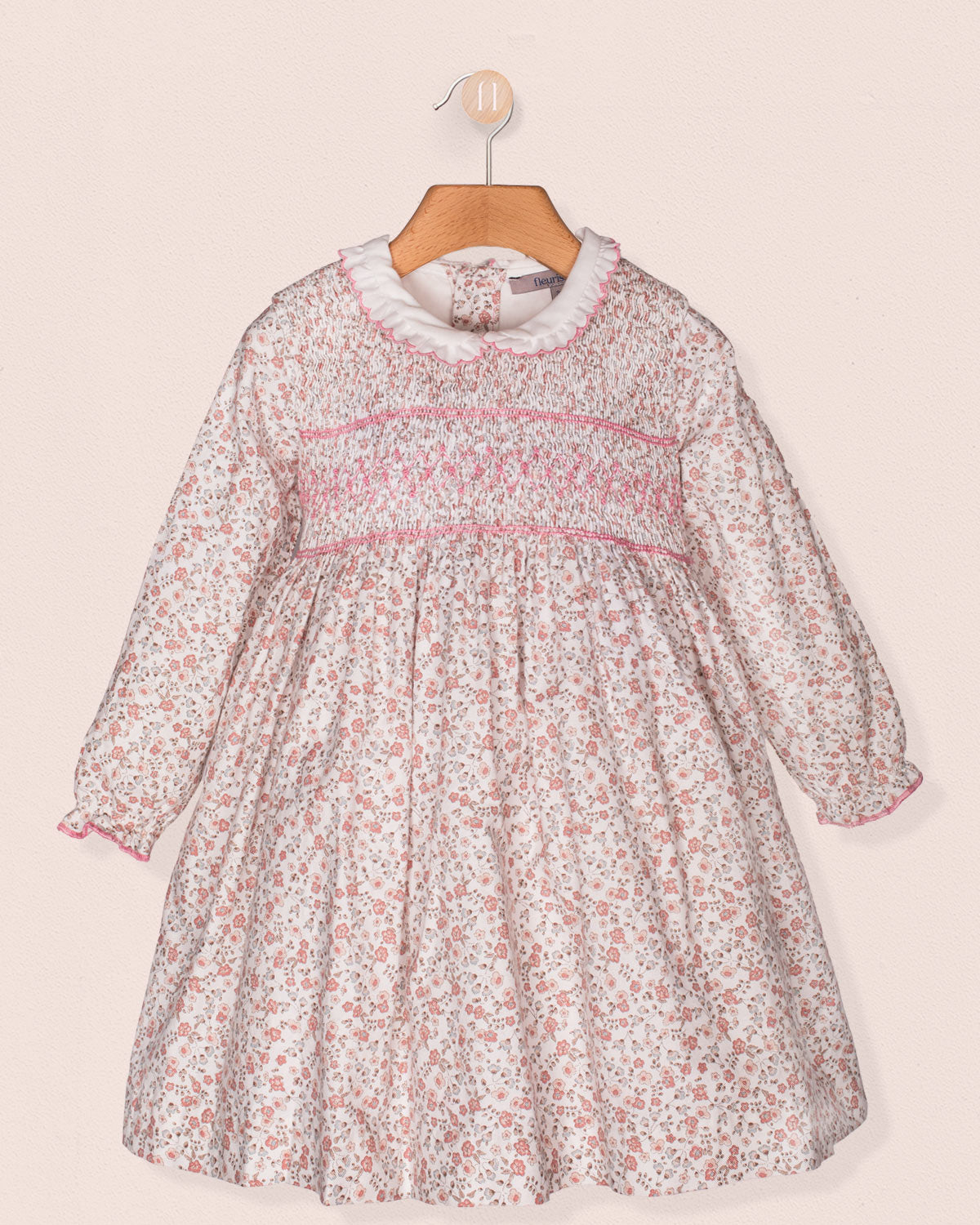 Front view of a baby girl's long-sleeve floral smocked dress with a white ruffle collar, pink hand-smocking across the bodice, and elasticated cuffs, hanging on a wooden hanger.