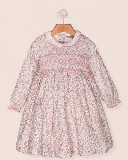 Front view of a baby girl's long-sleeve floral smocked dress with a white ruffle collar, pink hand-smocking across the bodice, and elasticated cuffs, hanging on a wooden hanger.