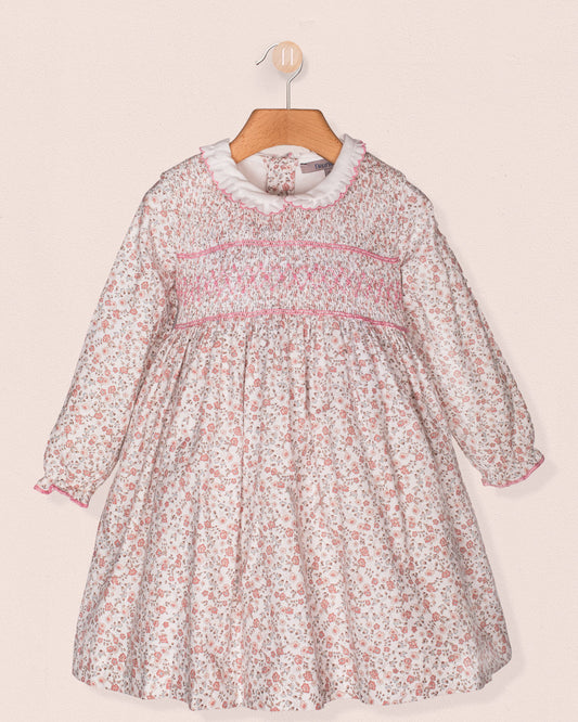 Front view of a baby girl's long-sleeve floral smocked dress with a white ruffle collar, pink hand-smocking across the bodice, and elasticated cuffs, hanging on a wooden hanger.