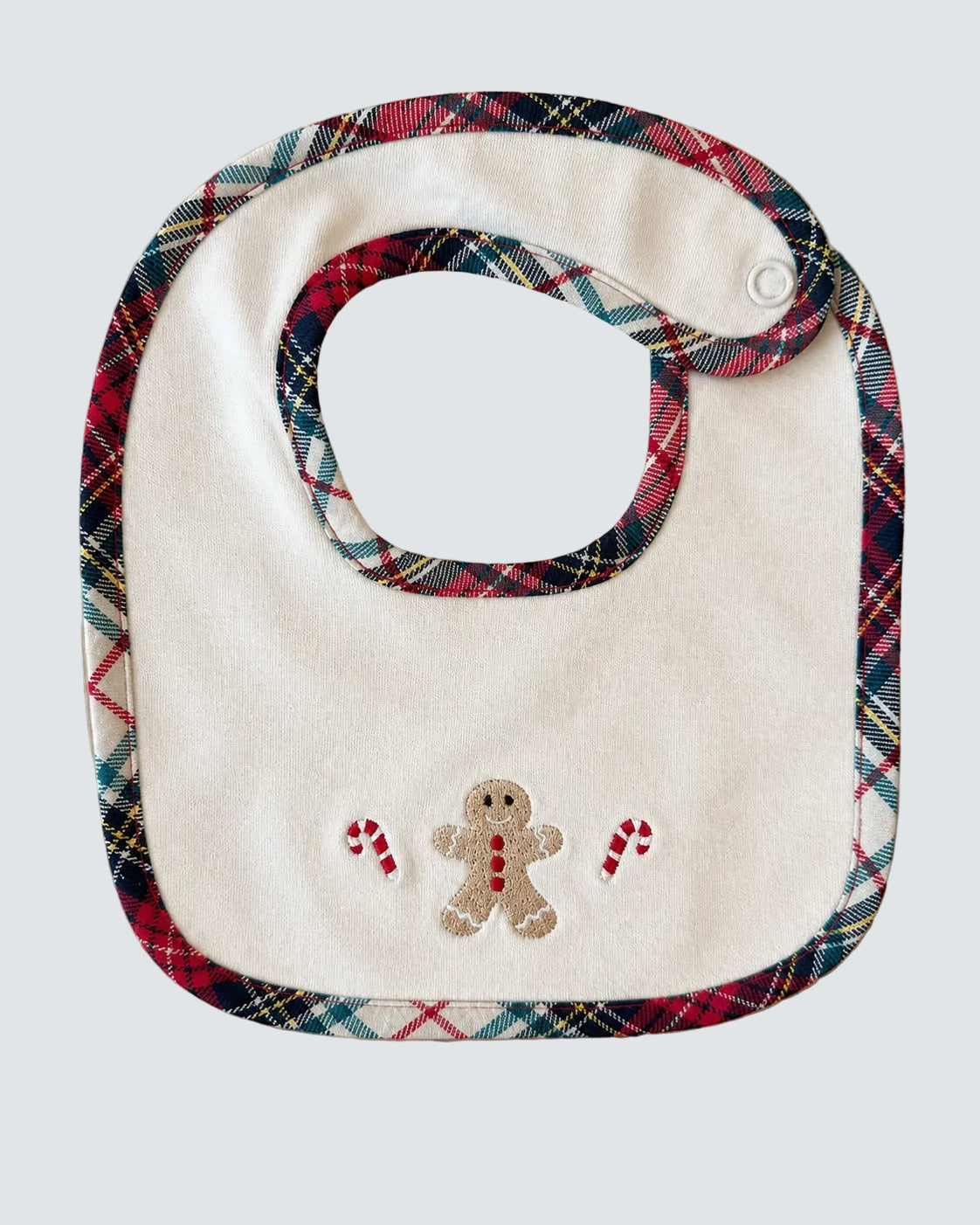 A baby bib with a cream-colored center and a red, green, and blue plaid border, featuring an embroidered gingerbread man and candy canes on the front, laid flat on a light wood background.