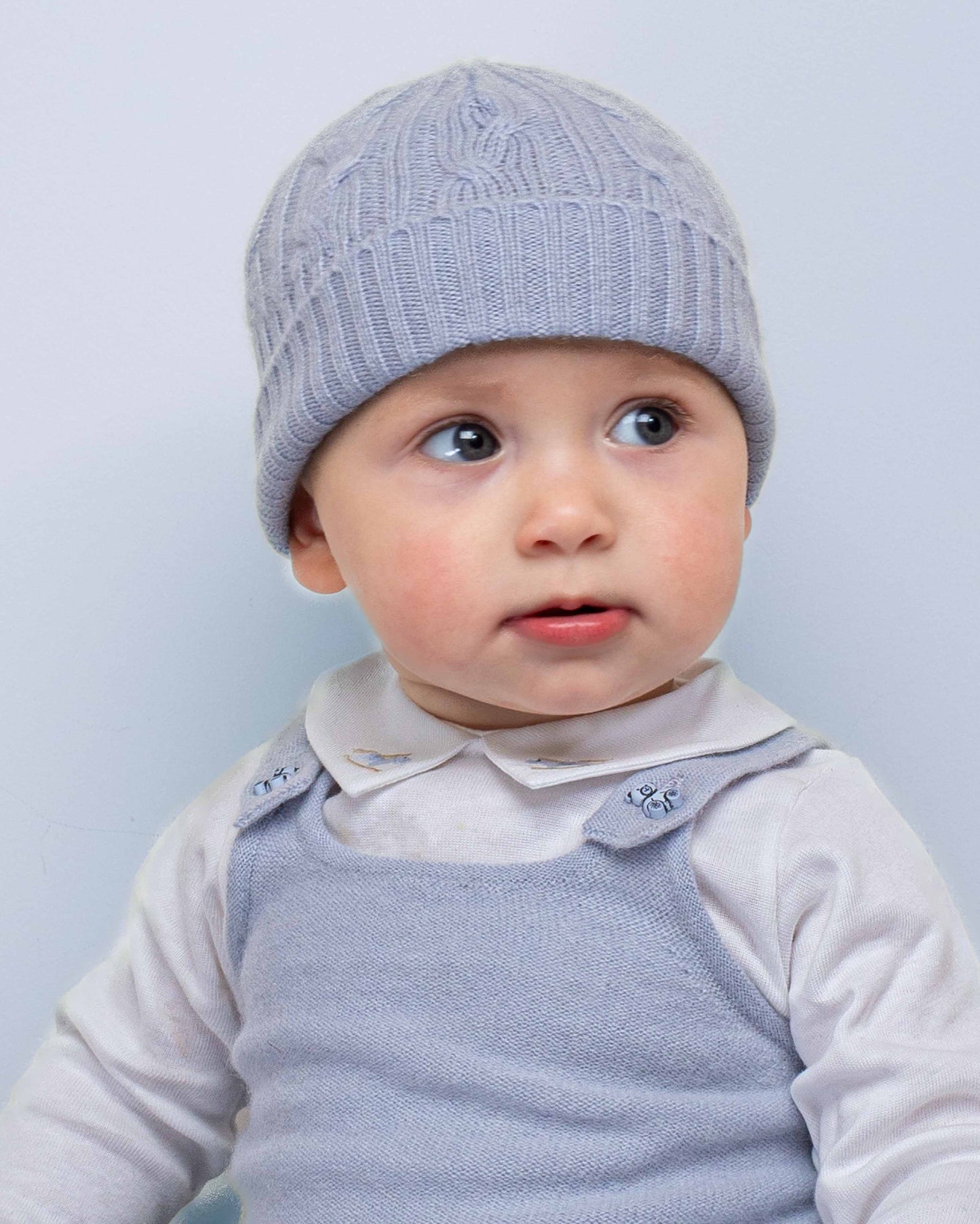 Toddler wearing a Cashmere Light Blue Cable Knit Hat, paired with a white top and blue knit overall, looking curiously to the side.