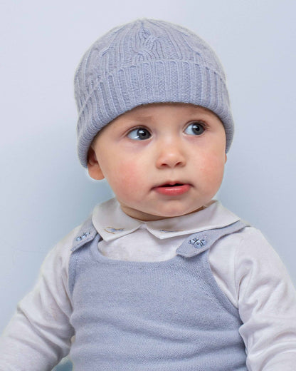 Toddler wearing a Cashmere Light Blue Cable Knit Hat, paired with a white top and blue knit overall, looking curiously to the side.