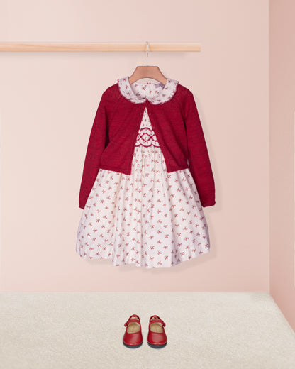 Red cardigan over a white dress with red floral pattern, paired with red shoes on a pink background.