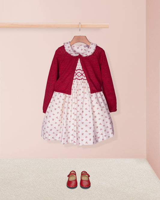 Red cardigan over a white dress with red floral pattern, paired with red shoes on a pink background.
