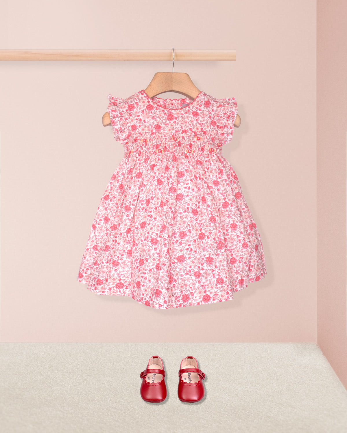 Pink floral dress on a hanger with red shoes on the floor against a pink wall.