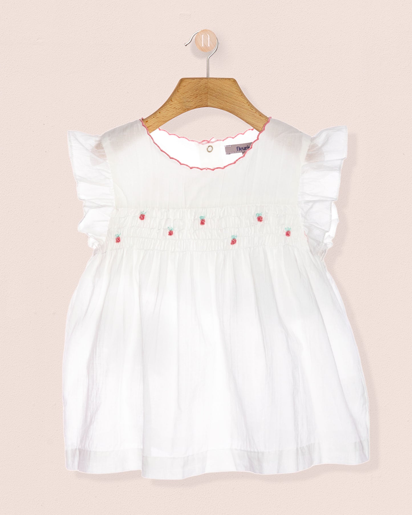 White tunic with strawberry designs on a hanger against a peach background.