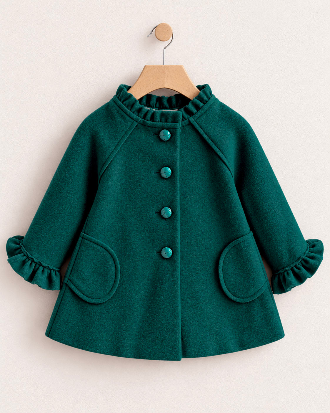 A dark hunter green children's coat with ruffled trim, button-front closure, and ruffled cuffs, hanging on a wooden hanger against a light beige wall.