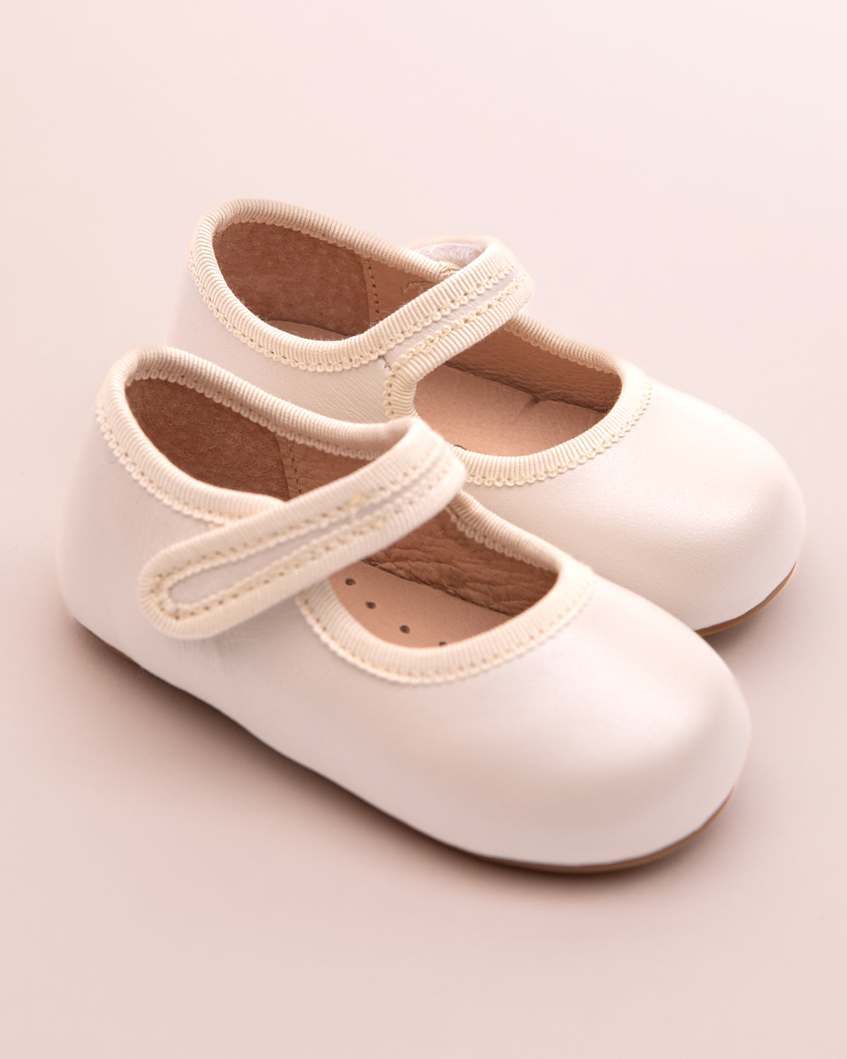 Pair of cream leather baby Mary Jane shoes with Velcro straps, placed on a blush pink background.
