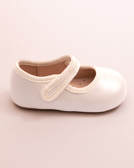 Side view of a cream leather baby Mary Jane shoe with a Velcro strap and soft sole, shown on a pink surface.
