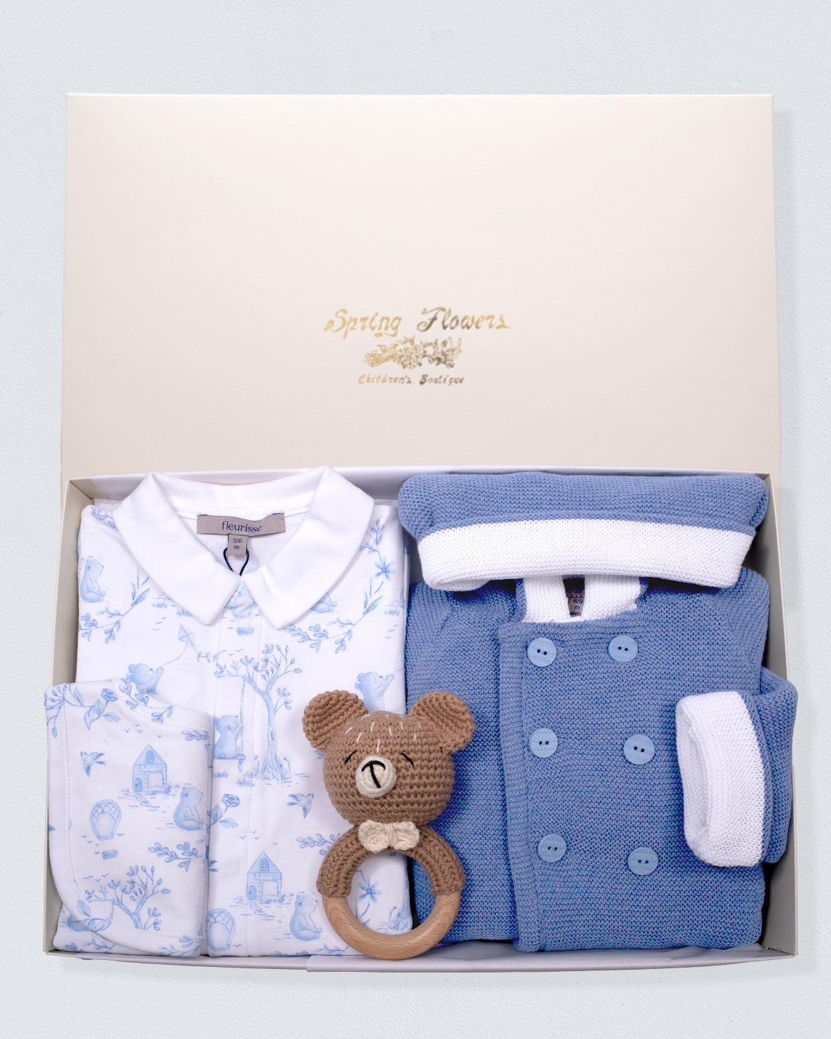 Baby clothing set with blue and white knit jacket outfit, white footie with blue patterns, and brown teddy bear rattle in a box.