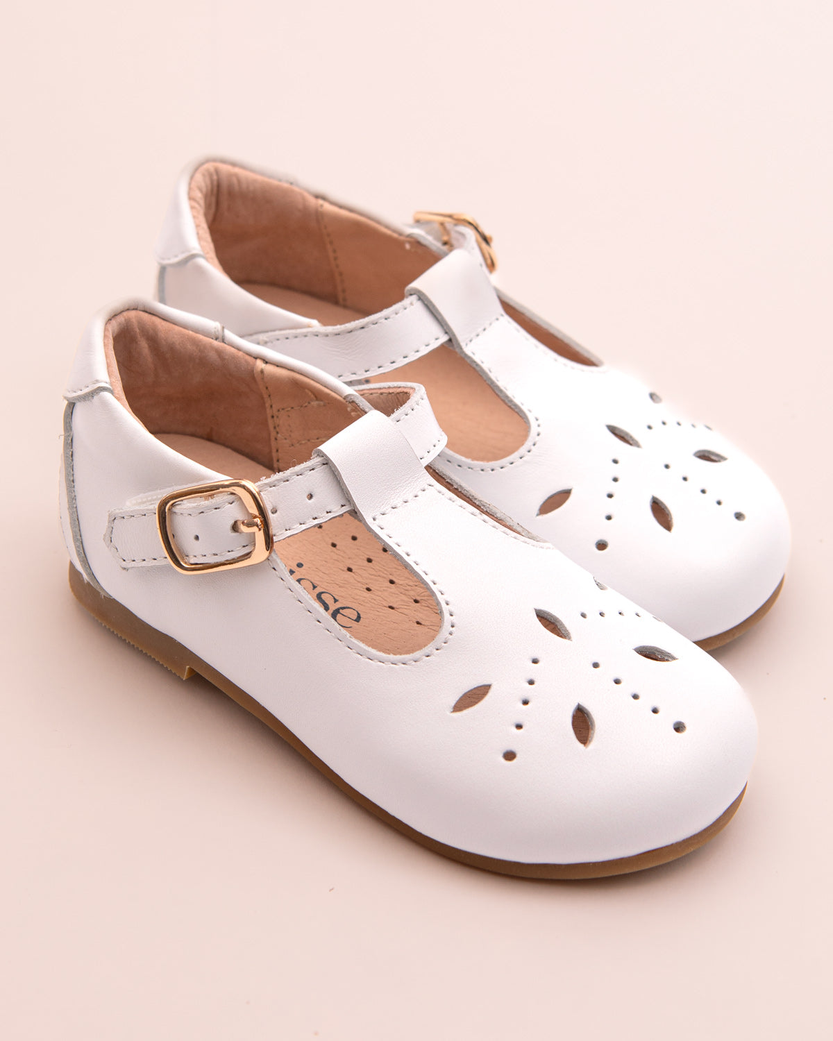 Pair of white leather T-bar shoes for toddlers with decorative cutout patterns, a velcro strap, and gold-tone buckle, displayed on a pale pink background.