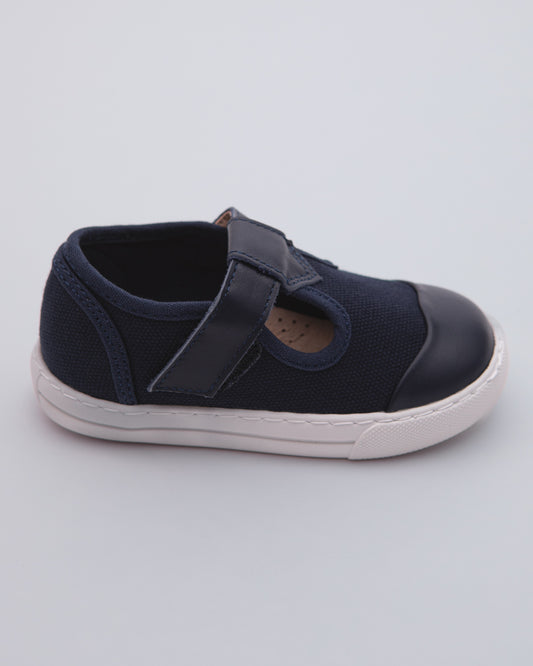 Side view of a navy blue toddler sneaker showing the white rubber sole, canvas body, leather toe cap, and velcro strap closure on a gray surface.