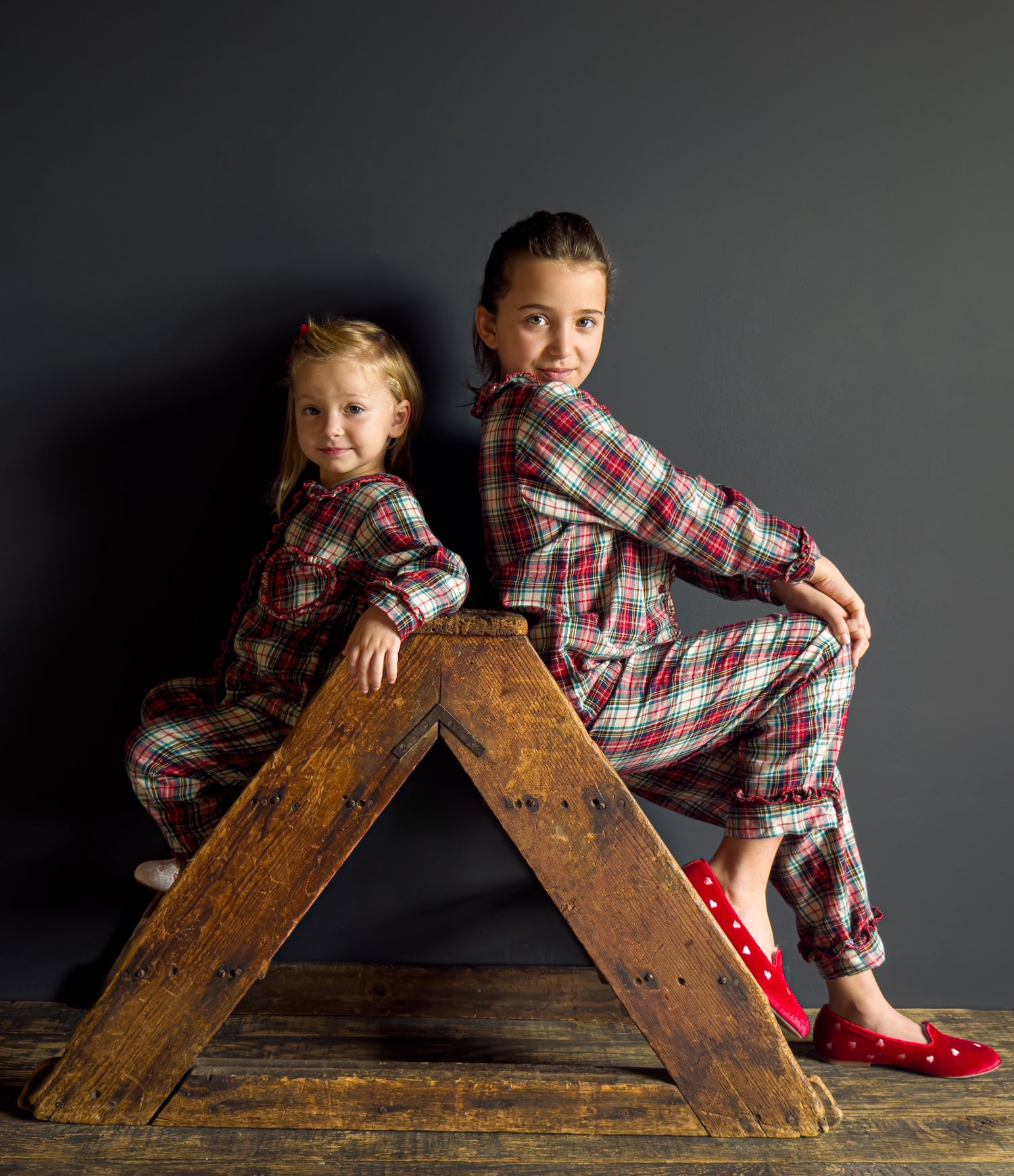 Two children in plaid pajamas sitting on a wooden ladder against a dark background
