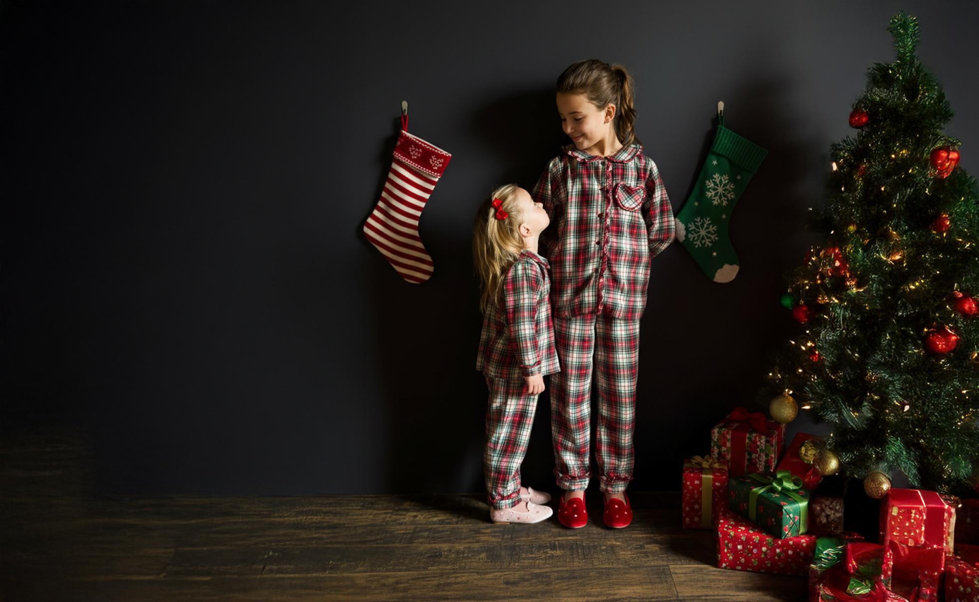 Two children in matching plaid pajamas standing in front of a Christmas tree with presents.