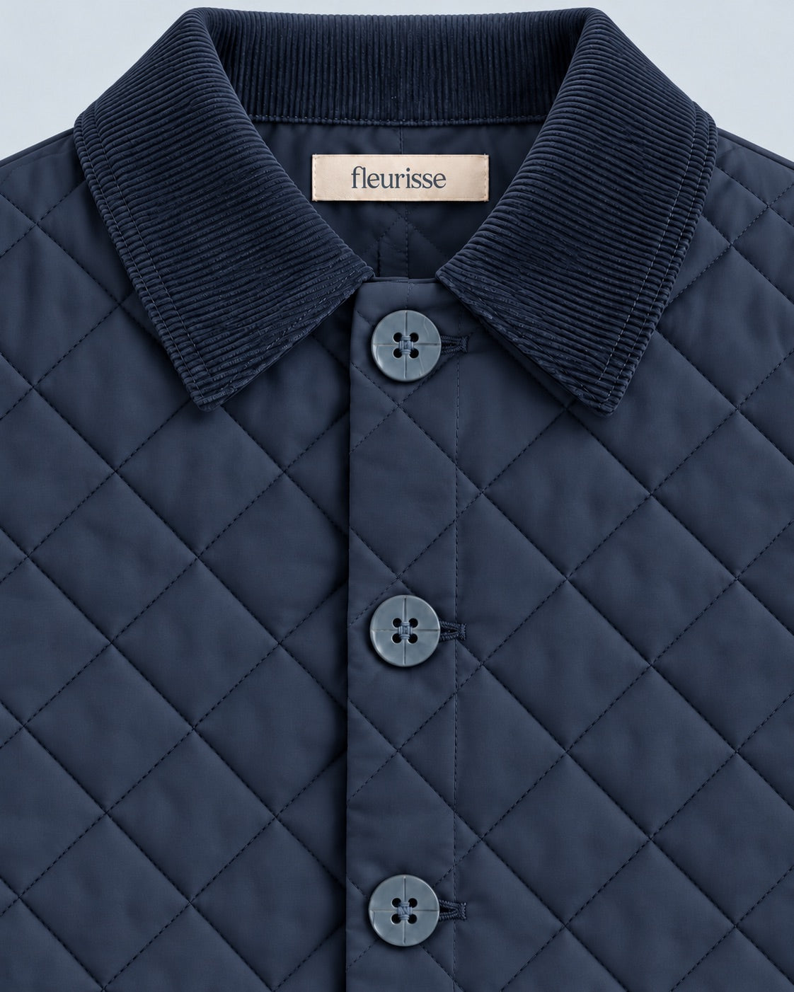 Close-up of a navy quilted collared jacket with a ribbed corduroy-style collar and matching buttons, showing a small Fleurisse label at the inner neck.