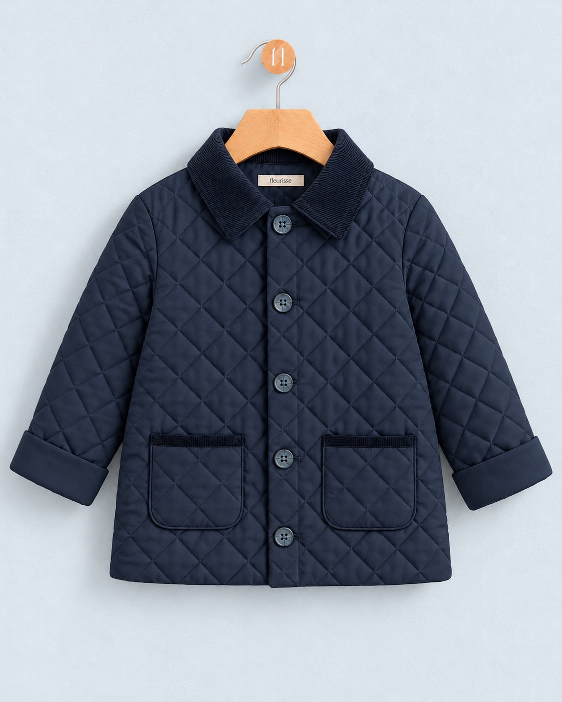 A navy blue quilted jacket for toddlers, featuring a corduroy collar, button-front closure, and two front patch pockets, hanging on a wooden hanger against a light gray background.