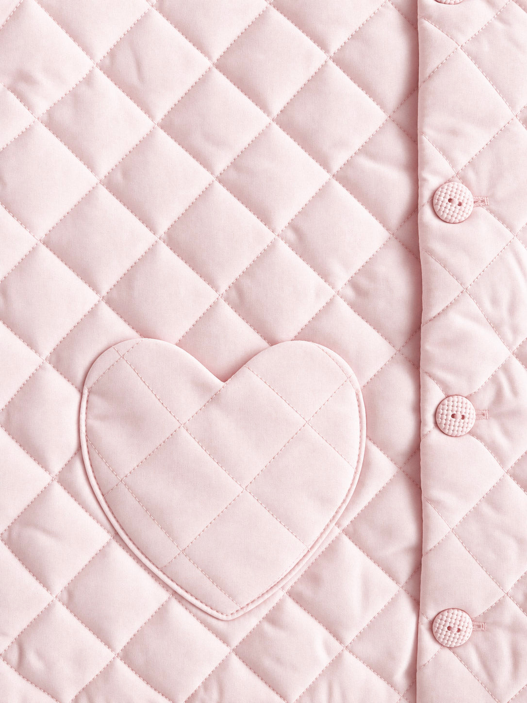 Close-up of a light pink quilted fabric with a diamond pattern, a heart-shaped patch pocket, and a buttoned edge.