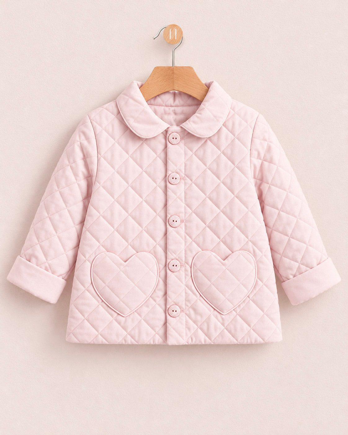 A light pink quilted baby jacket with a Peter Pan collar, button front, and heart-shaped pockets, hanging on a wooden hanger against a solid pale pink background.