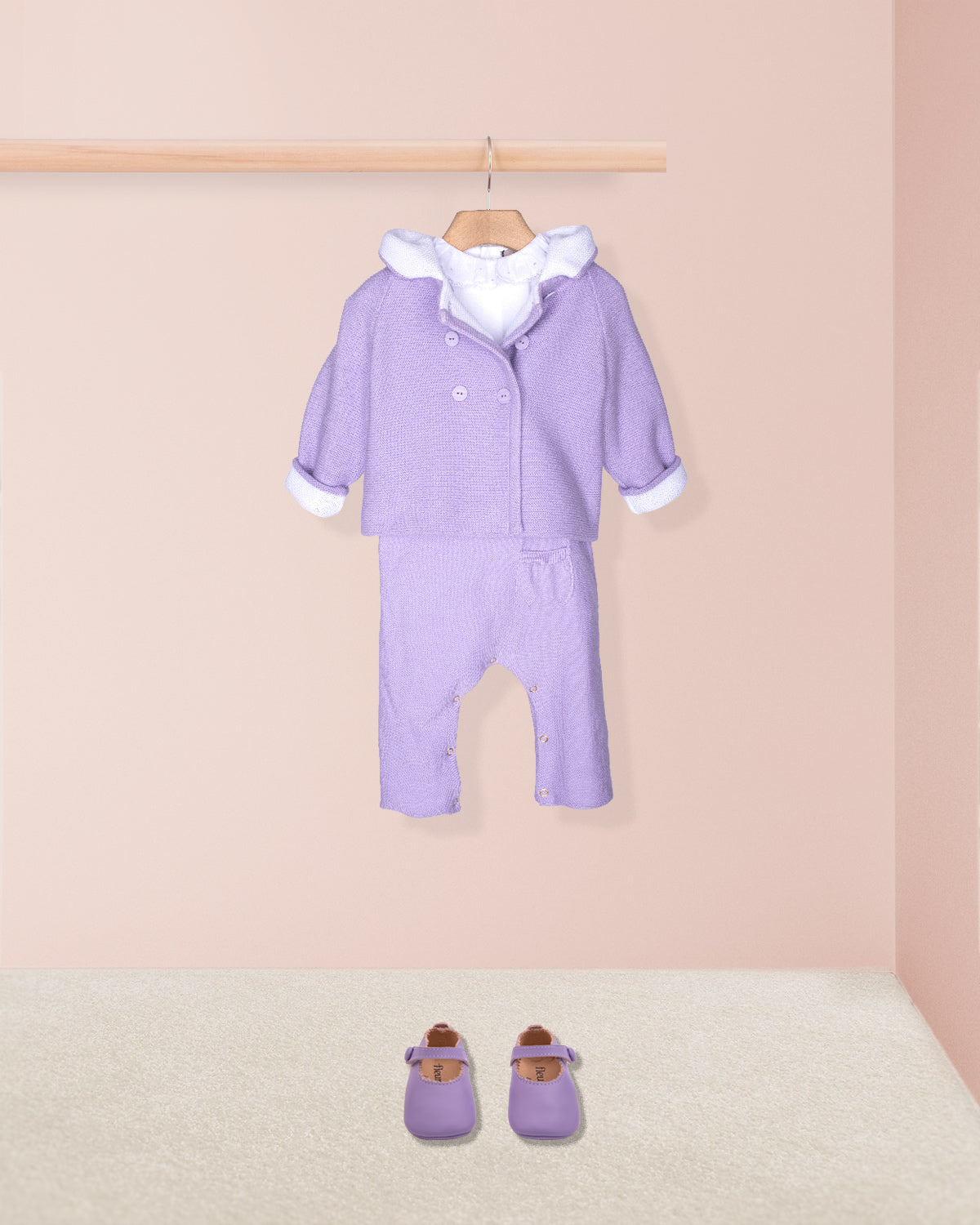 Purple baby outfit with white trim on a wooden hanger against a pink wall.