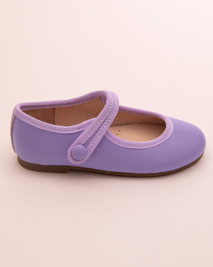 Side view of a single lavender leather Mary-Jane shoe with velcro strap, rubber sole, and tonal grosgrain trim on a light pink surface.