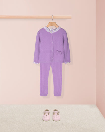 Purple children's outfit with a matching hat on a hanger against a pink wall.