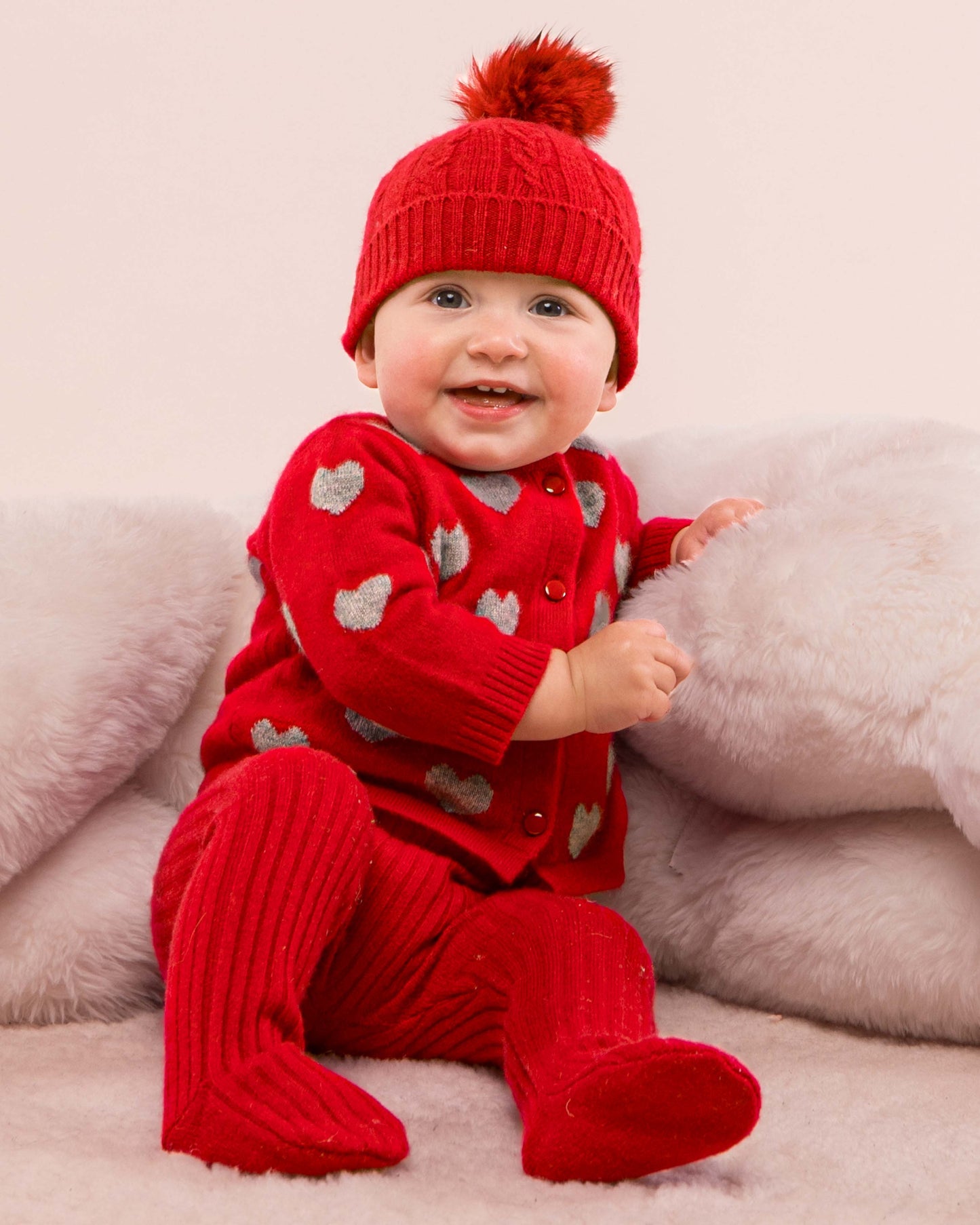 Baby model wearing the Madeleine Red Hearts Cashmere Cardigan sitting down showing the front of the sweater, along with a matching cashmere leggings and a hat 