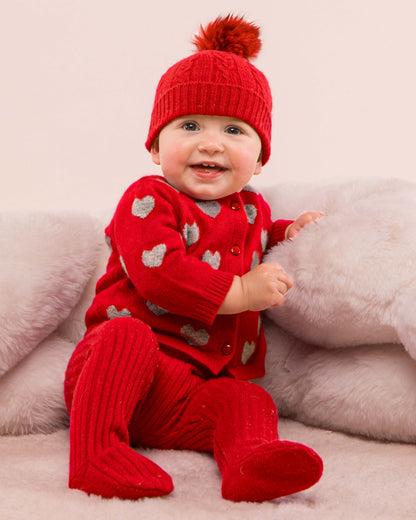 Baby model wearing the Madeleine Red Hearts Cashmere Cardigan sitting down showing the front of the sweater, along with a matching cashmere leggings and a hat 