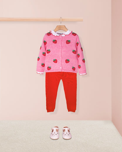 Pink sweater with strawberry pattern and red pants on a hanger against a pink background