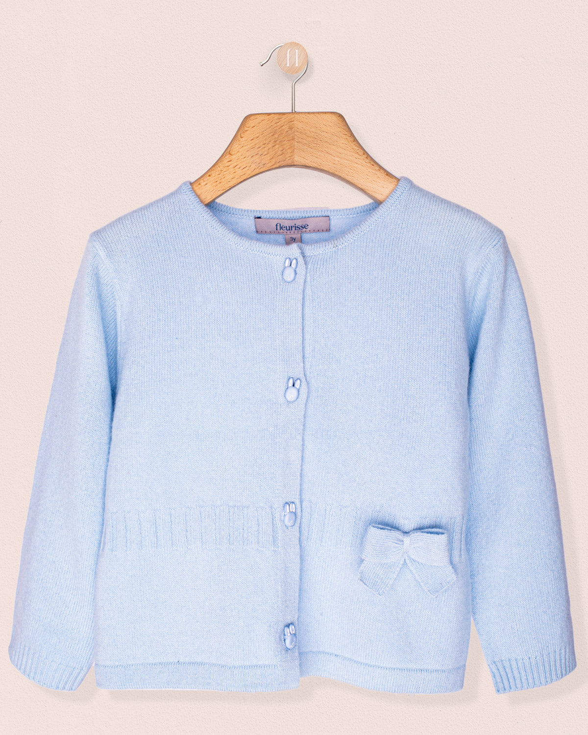 Light blue cardigan with a bow and bunny buttons, on a wooden hanger against a beige background