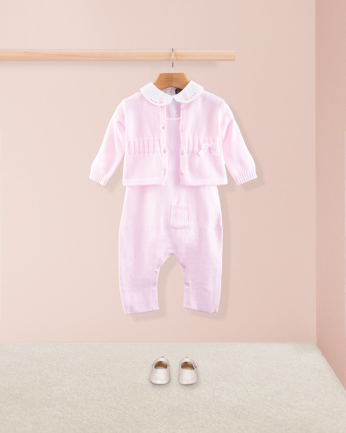 Pink cotton knit baby overall layered with a matching pink knit cardigan and paired with pink baby shoes on a beige carpet