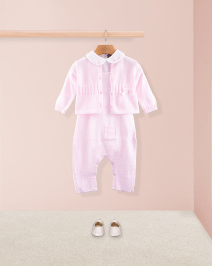 Pink cotton knit baby overall layered with a matching pink knit cardigan and paired with pink baby shoes on a beige carpet
