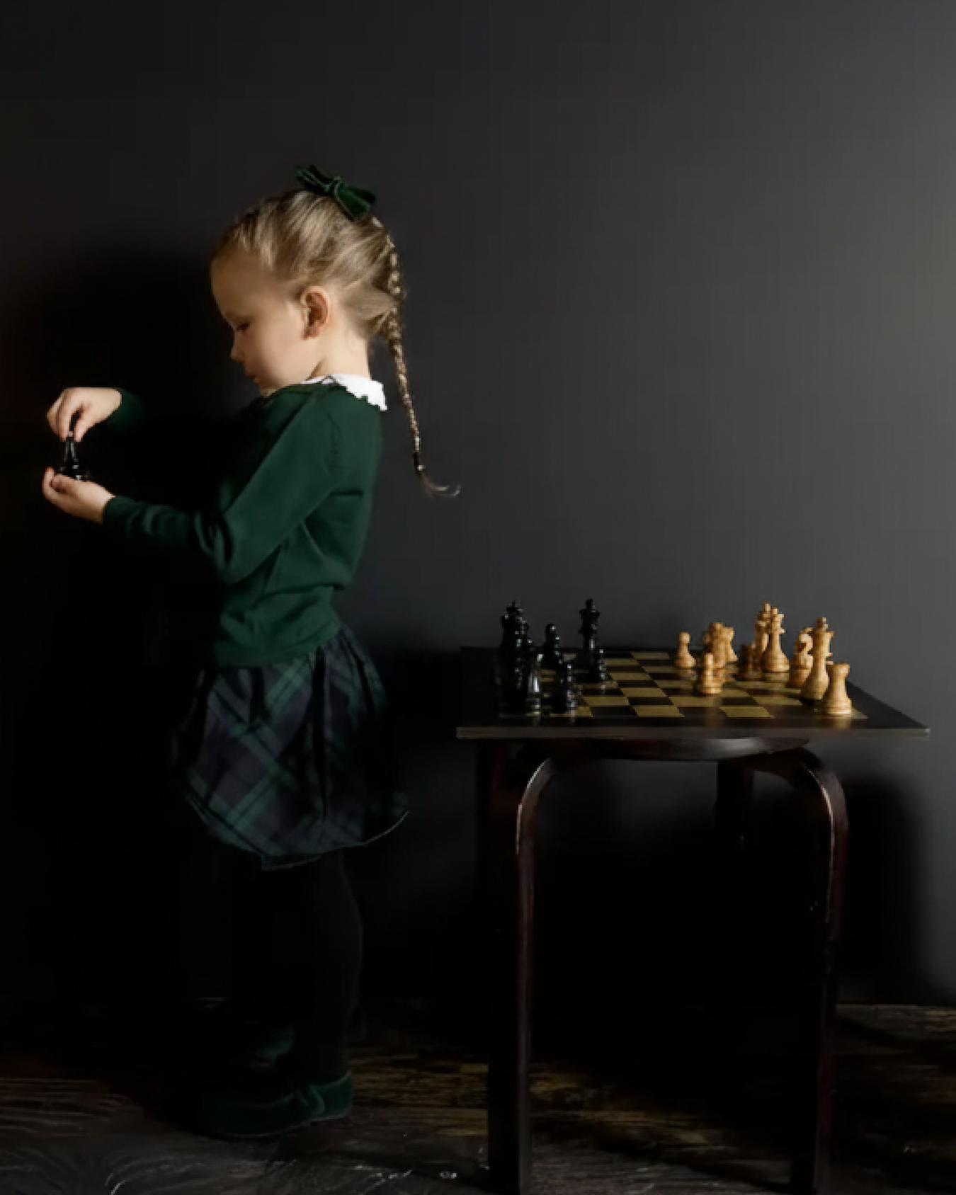 Young girl playing with a chess set on a small table against a dark wall.