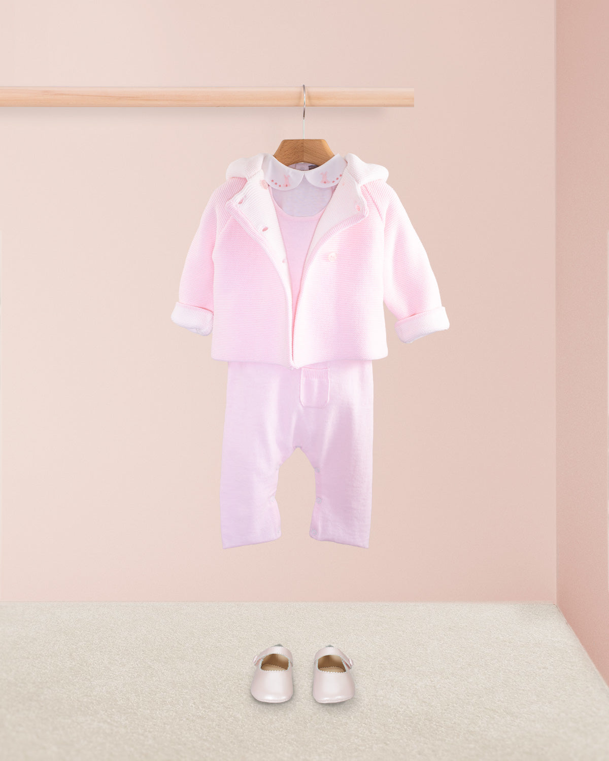 Pink cotton knit baby overall layered with a pink knit hooded jacket over a white embroidered collar onesie and paired with pink baby shoes on a beige carpet