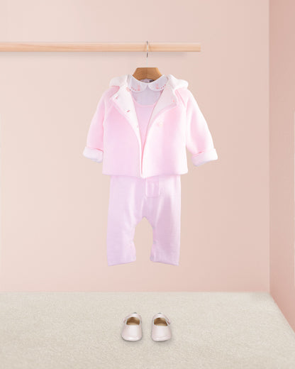 Pink cotton knit baby overall layered with a pink knit hooded jacket over a white embroidered collar onesie and paired with pink baby shoes on a beige carpet