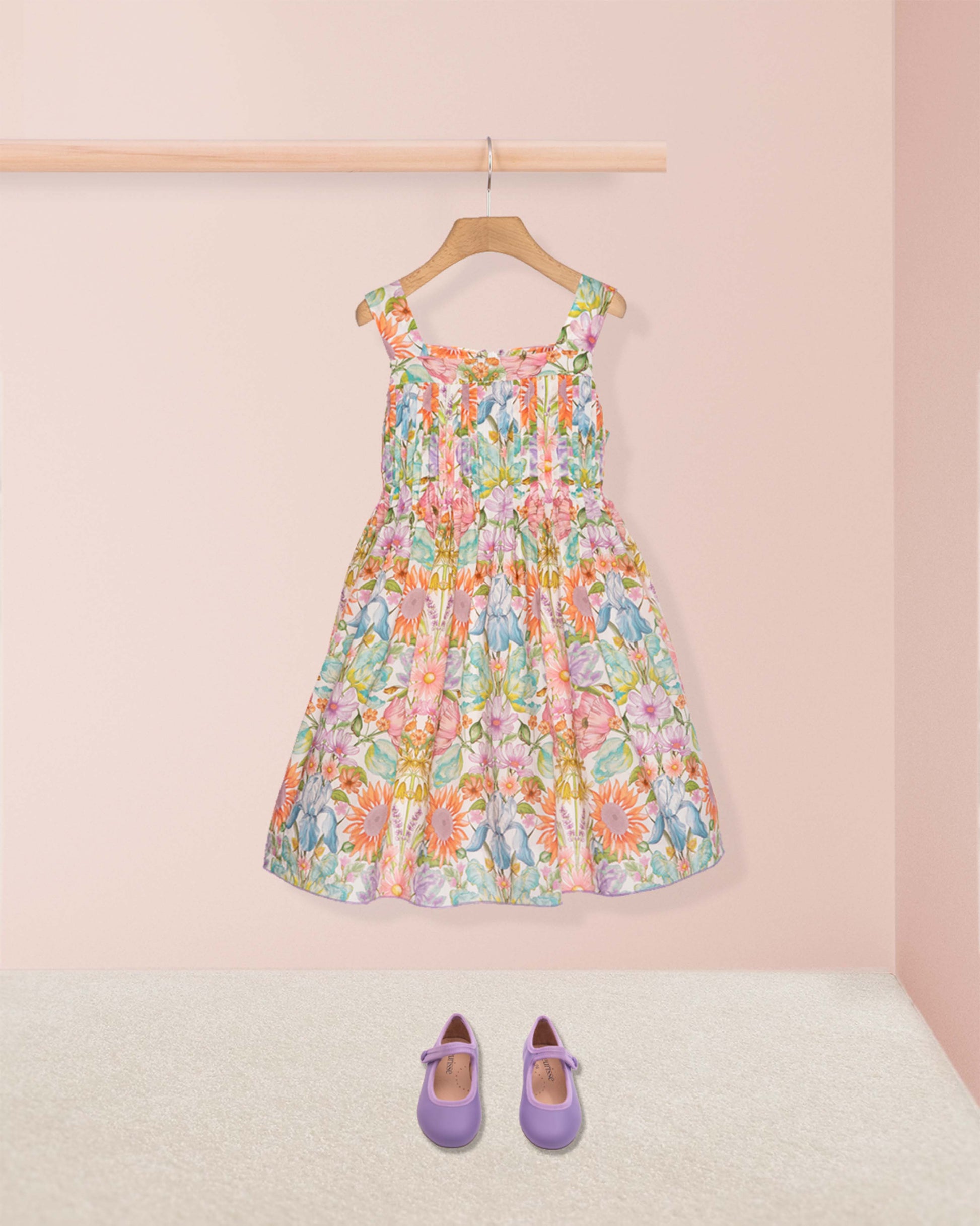 Colorful floral dress on a hanger with purple shoes on the floor against a pink wall.