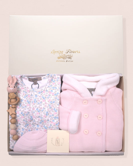Baby gift set with pink outfit, floral onesie, and baby hat in a box on a light pink background.
