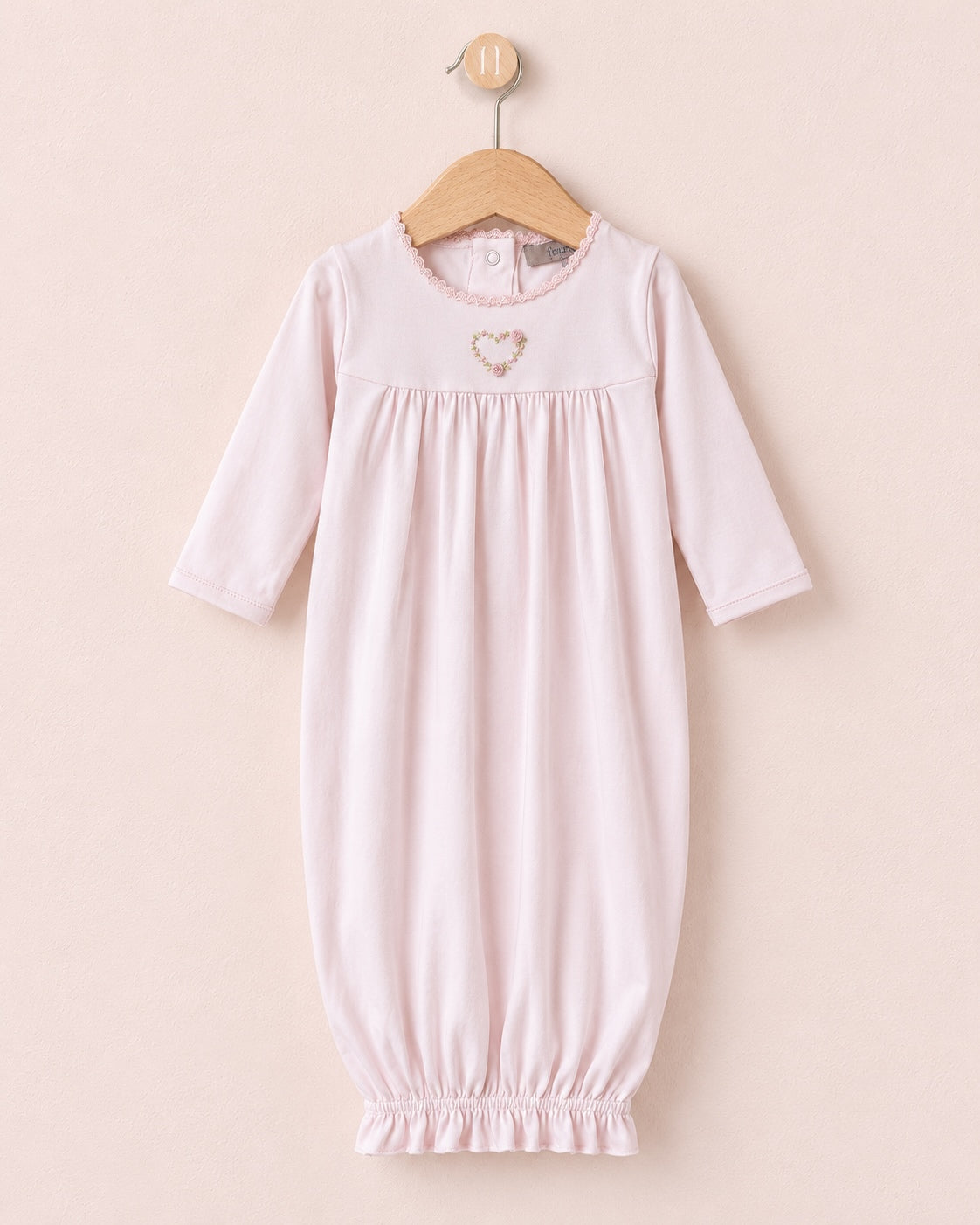 A light pink baby lounging gown with three-quarter sleeves and an elasticated hem, hanging on a wooden hanger against a soft beige background. It features a small embroidered heart on the chest.