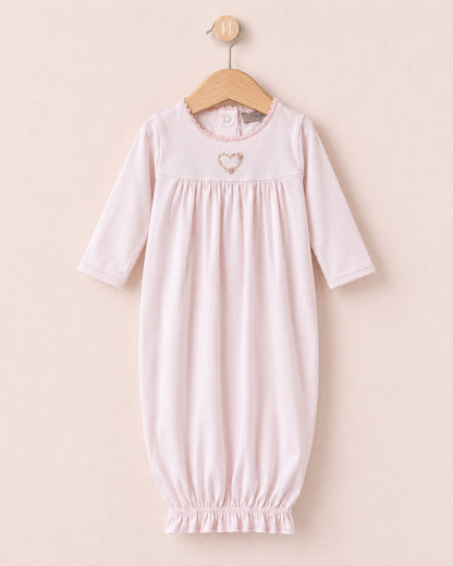 A light pink baby lounging gown with three-quarter sleeves and an elasticated hem, hanging on a wooden hanger against a soft beige background. It features a small embroidered heart on the chest.