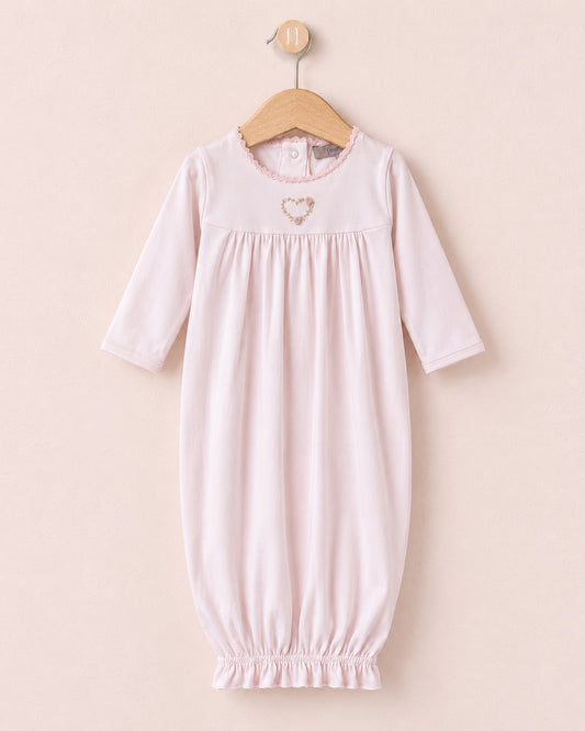 A light pink baby lounging gown with three-quarter sleeves and an elasticated hem, hanging on a wooden hanger against a soft beige background. It features a small embroidered heart on the chest.