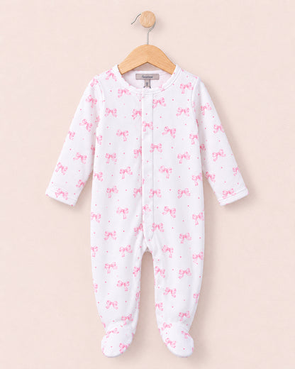 A baby footed sleeper in white with a pink bow and dot pattern, hanging on a wooden hanger against a solid pale pink background.